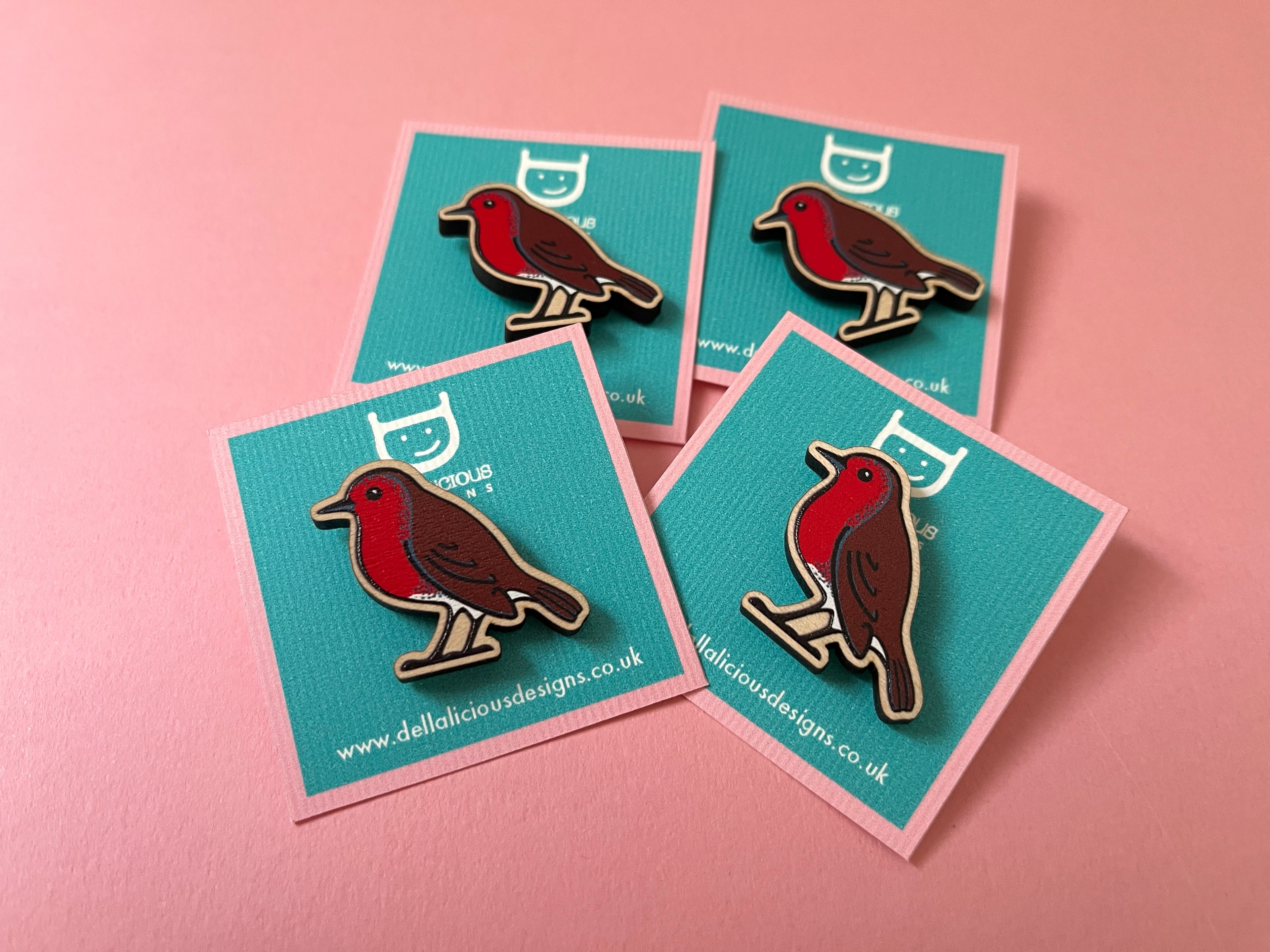 Four enamel pins of a robin design on blue cards with a pink background