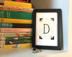 A black framed picture with a white background and a large capital letter D in black.
