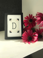A black picture frame with a white card inside. The card has a large black letter 'D' in the center and four red corners with the numbers '6' and '9' on them. The frame is sitting on a white surface with three pink flowers behind it.