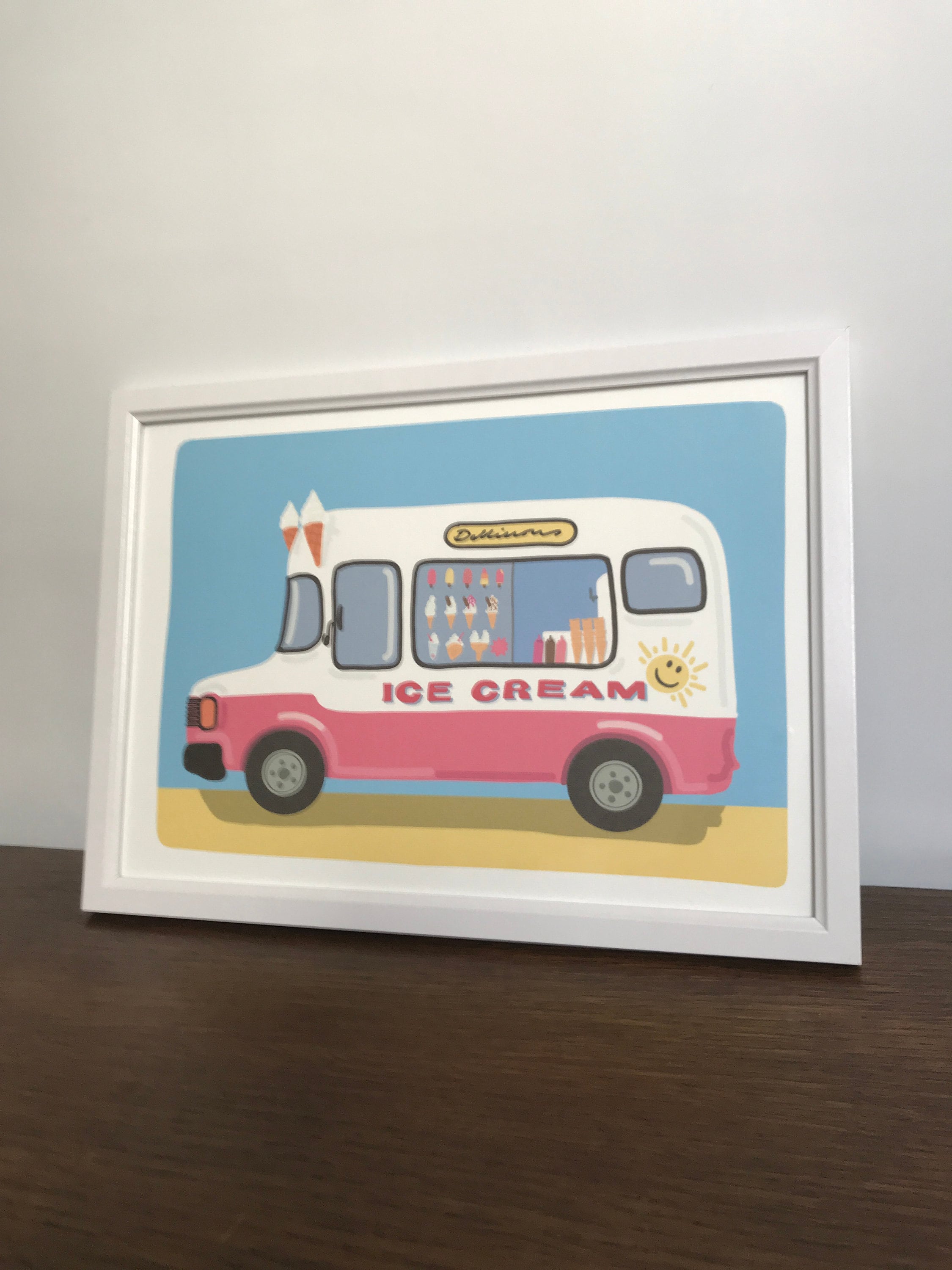 A framed illustration of a pink and white ice cream truck on a light blue background. The truck has the words 'Delicious' and 'Ice Cream' on it.  The ice cream truck is surrounded by a yellow sandy beach. The illustration is in a white frame. The image is a whimsical and colorful depiction of a classic summer treat.