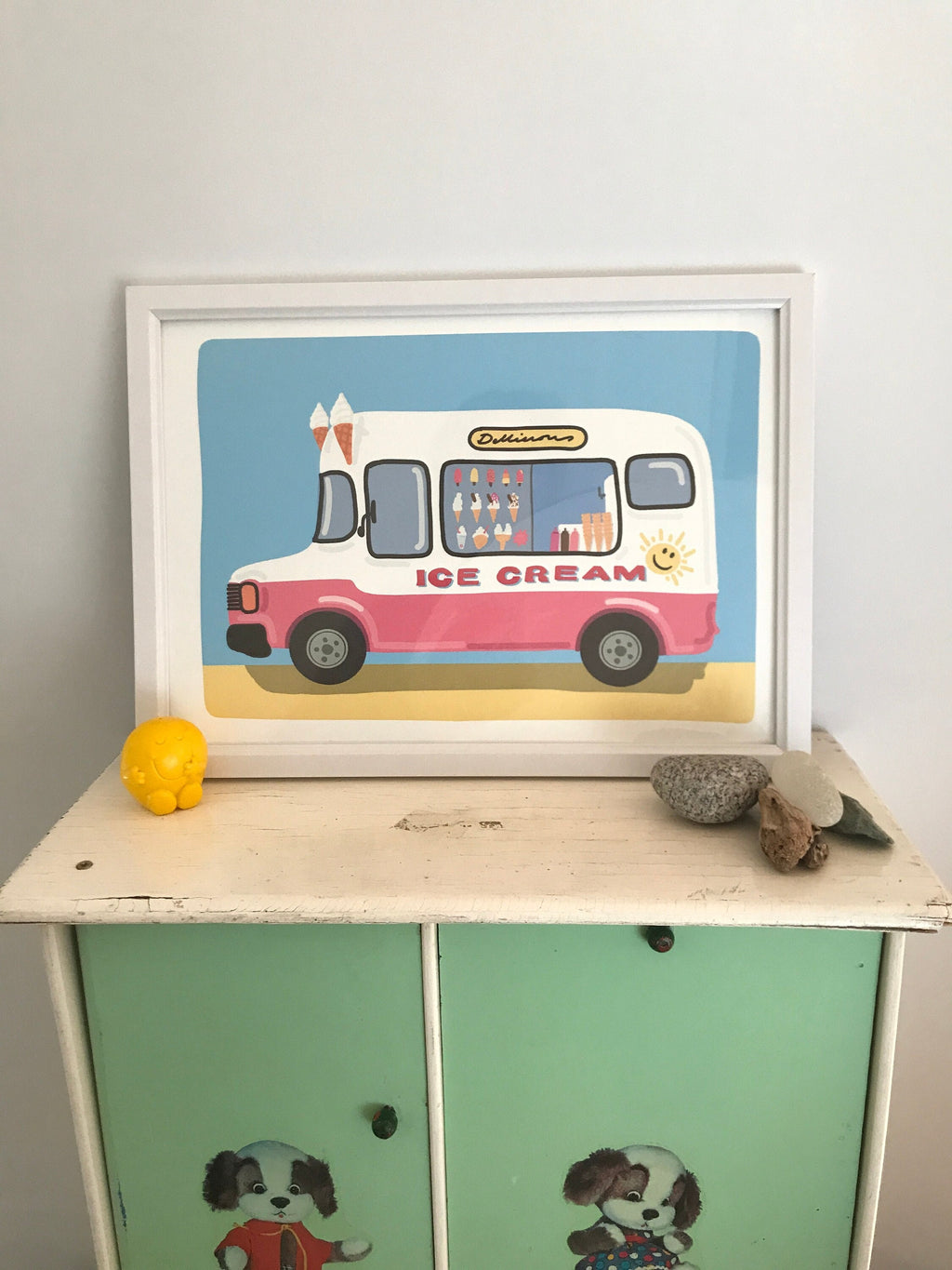 A framed illustration of a pink ice cream truck on a light blue background. The truck has the words 'Delicious' and 'Ice Cream' on it.  The window displays various ice cream cones and other treats. The truck has a smiling sun decal on the side. The illustration is displayed on a light green cabinet with painted doors