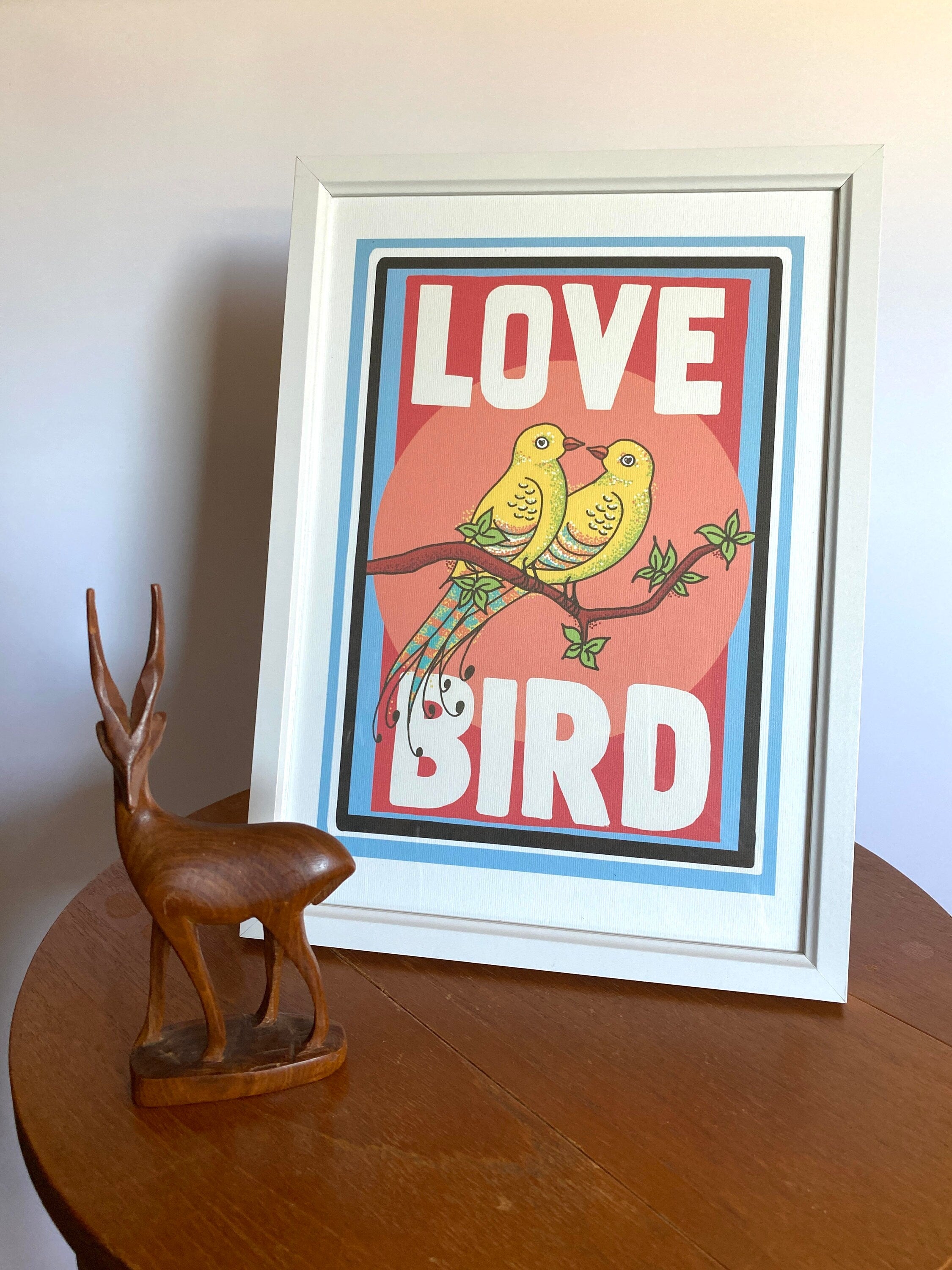 A framed print sits on a wooden surface against a white background next to a wooden deer. The print has a white border and features two yellow birds with their beaks touching sitting on a branch. the background behind the birds is pink. Large white text reads Love Bird all written in upper case letters.