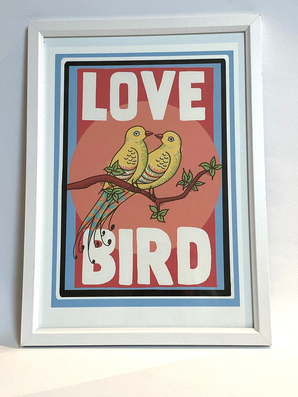 A framed print against a white background. The print has a white border and features two yellow birds with their beaks touching sitting on a branch. the background behind the birds is pink. Large white text reads Love Bird all written in upper case letters.
