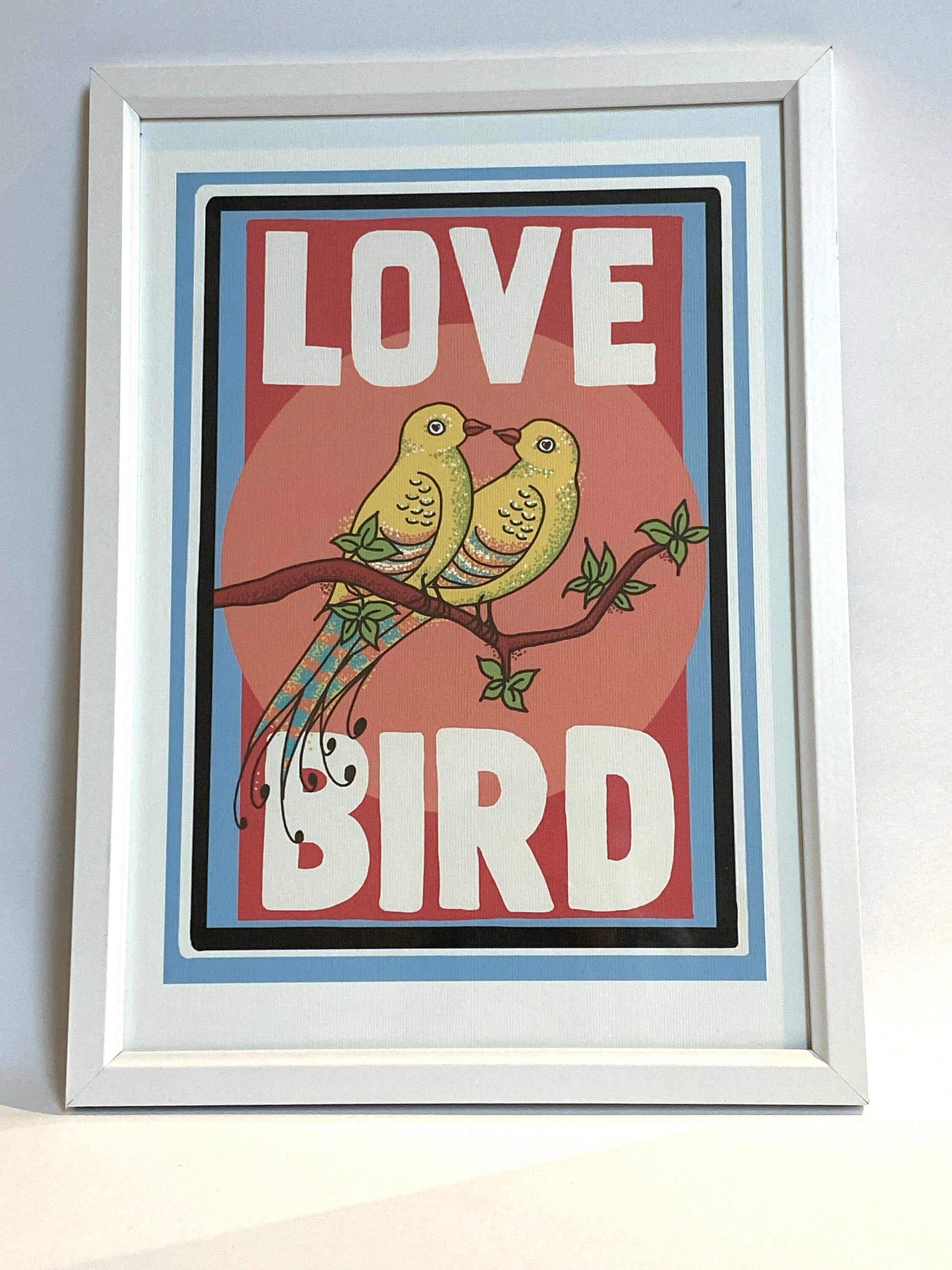 A framed print against a white background. The print has a white border and features two yellow birds with their beaks touching sitting on a branch. the background behind the birds is pink. Large white text reads Love Bird all written in upper case letters.