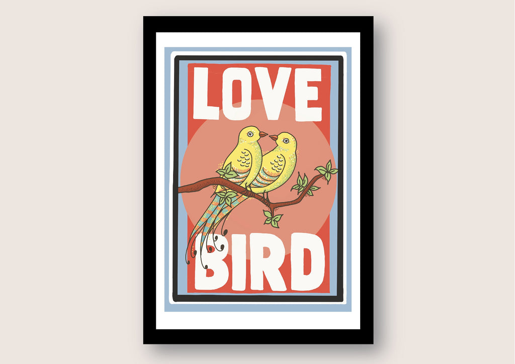 A black framed print against a white background. The print has a white border and features two yellow birds with their beaks touching sitting on a branch. the background behind the birds is pink. Large white text reads Love Bird all written in upper case letters.