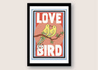 A black framed print against a white background. The print has a white border and features two yellow birds with their beaks touching sitting on a branch. the background behind the birds is pink. Large white text reads Love Bird all written in upper case letters.