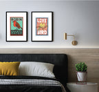 Two black framed prints on a grey wall above a bed with big pillows . One print has a white border and features two yellow birds with their beaks touching sitting on a branch. the background behind the birds is pink. Large white text reads Love Bird all written in upper case letters. The other print shows a Robin.