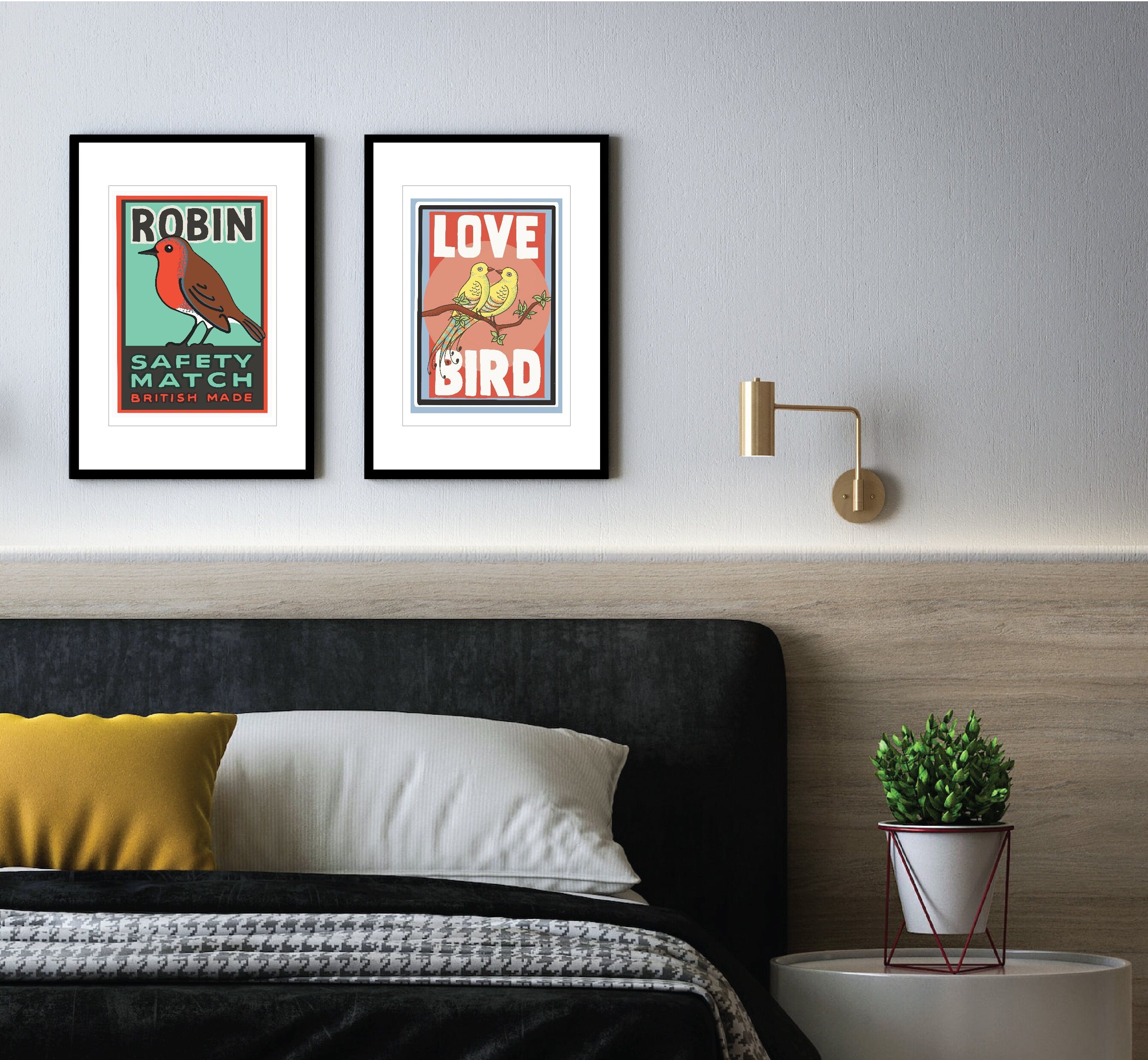 Two black framed prints on a grey wall above a bed with big pillows . One print has a white border and features two yellow birds with their beaks touching sitting on a branch. the background behind the birds is pink. Large white text reads Love Bird all written in upper case letters. The other print shows a Robin.