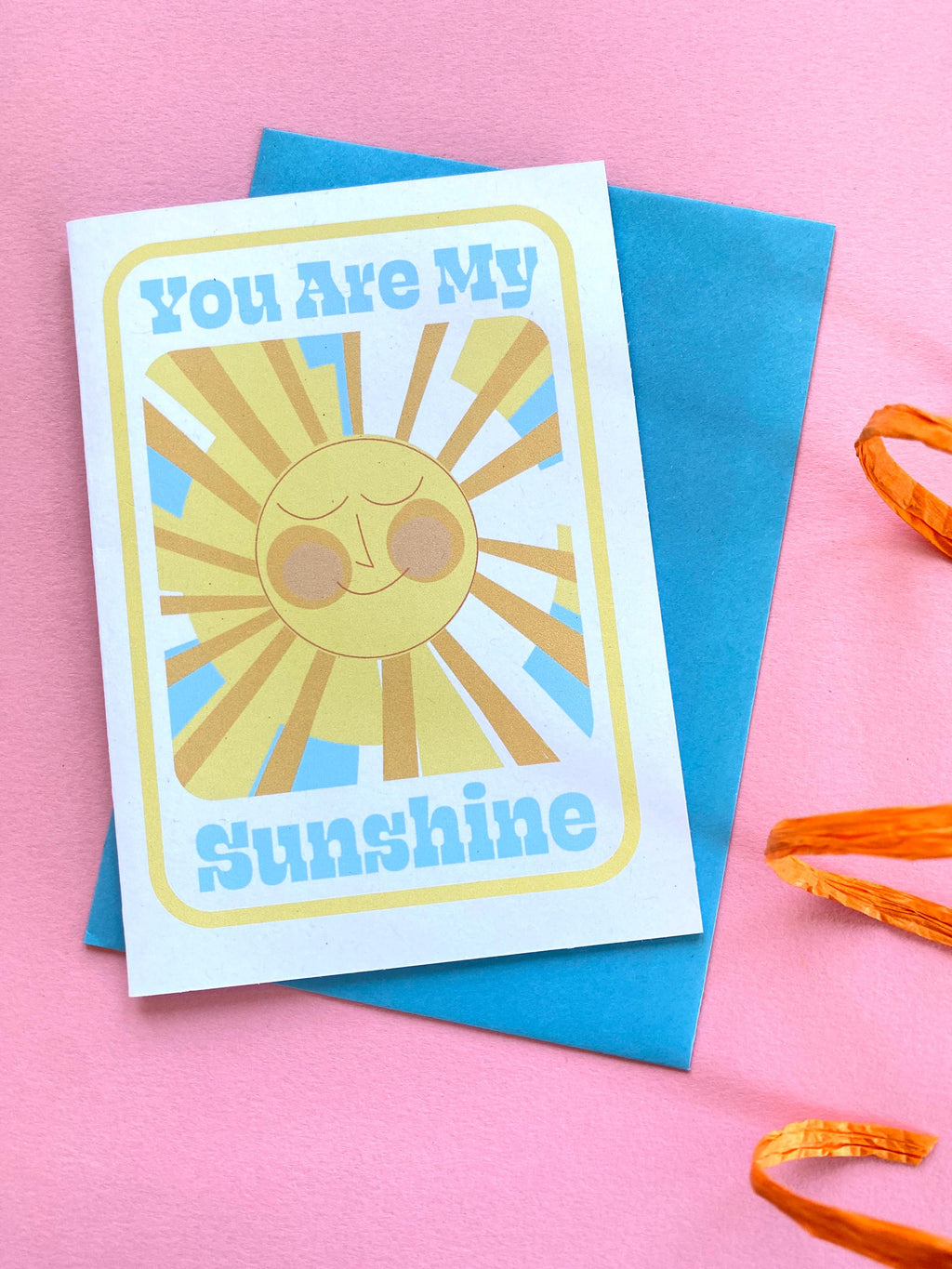 A birthday card sits on a blue envelope on a pink background with an orange ribbon at the side. The Birthday card is white with smiley illustrated sun in yellows and oranges with rays shining out from it. The text on the card reads You are my sunshine in light blue.