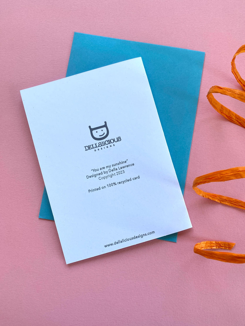 The back of a birthday card lays on a blue envelope on a pink background with orange ribbon at the side.