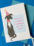 A close up of a birthday card sits on a pale blue envelope on a rich blue background with a curly ribbon. The birthday card has a illustration of a seated black greyhound with a white chest and stripy cone shaped birthday hat. The message on the front reads - Wishing you the happiest of Birthdays filled with treats. The text is in pink on a very light blue background.