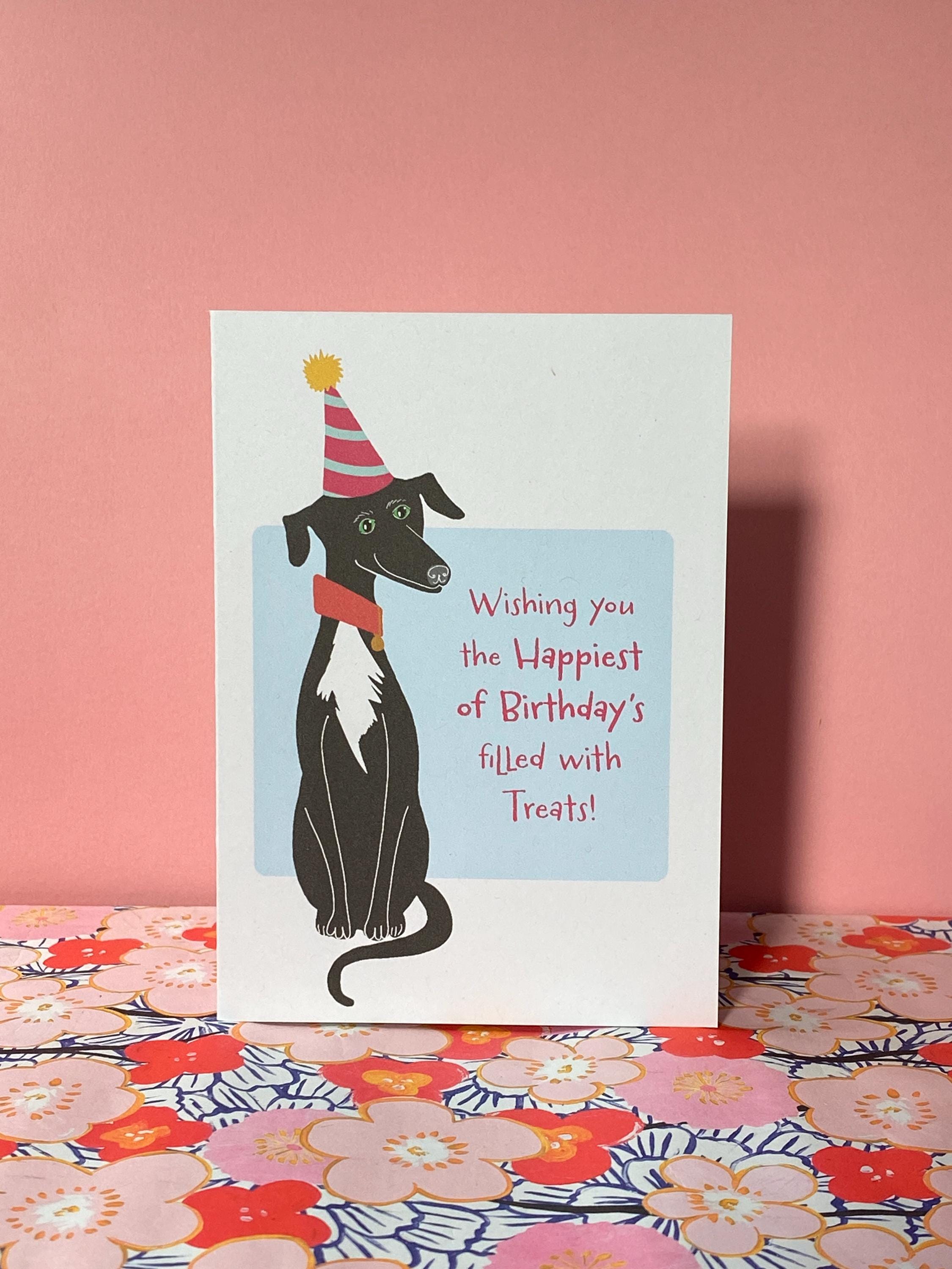A birthday card sits pink floral shelf against a pink background. The birthday card has a illustration of a seated black greyhound with a white chest and stripy cone shaped birthday hat. The message on the front reads - Wishing you the happiest of Birthdays filled with treats. The text is in pink on a very light blue background.