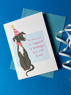 A birthday card sits on a pale blue envelope on a rich blue background with a curly ribbon. The birthday card has a illustration of a seated black greyhound with a white chest and stripy cone shaped birthday hat. The message on the front reads - Wishing you the happiest of Birthdays filled with treats. The text is in pink on a very light blue background.