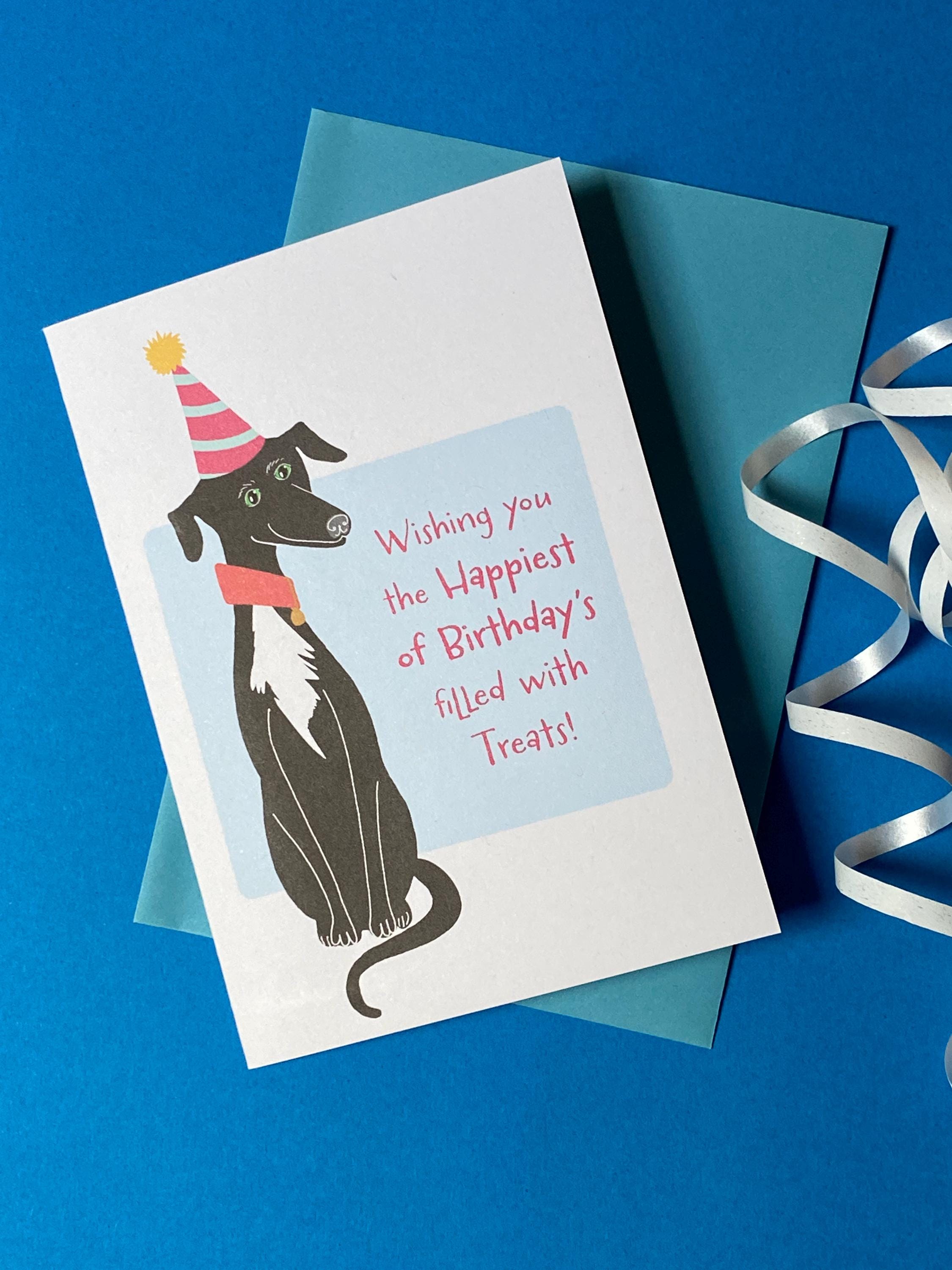A birthday card sits on a pale blue envelope on a rich blue background with a curly ribbon. The birthday card has a illustration of a seated black greyhound with a white chest and stripy cone shaped birthday hat. The message on the front reads - Wishing you the happiest of Birthdays filled with treats. The text is in pink on a very light blue background.