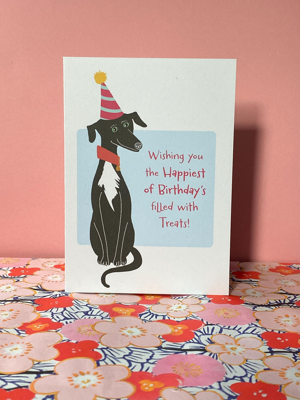 A birthday card sits pink floral shelf against a pink background. The birthday card has a illustration of a seated black greyhound with a white chest and stripy cone shaped birthday hat. The message on the front reads - Wishing you the happiest of Birthdays filled with treats. The text is in pink on a very light blue background.