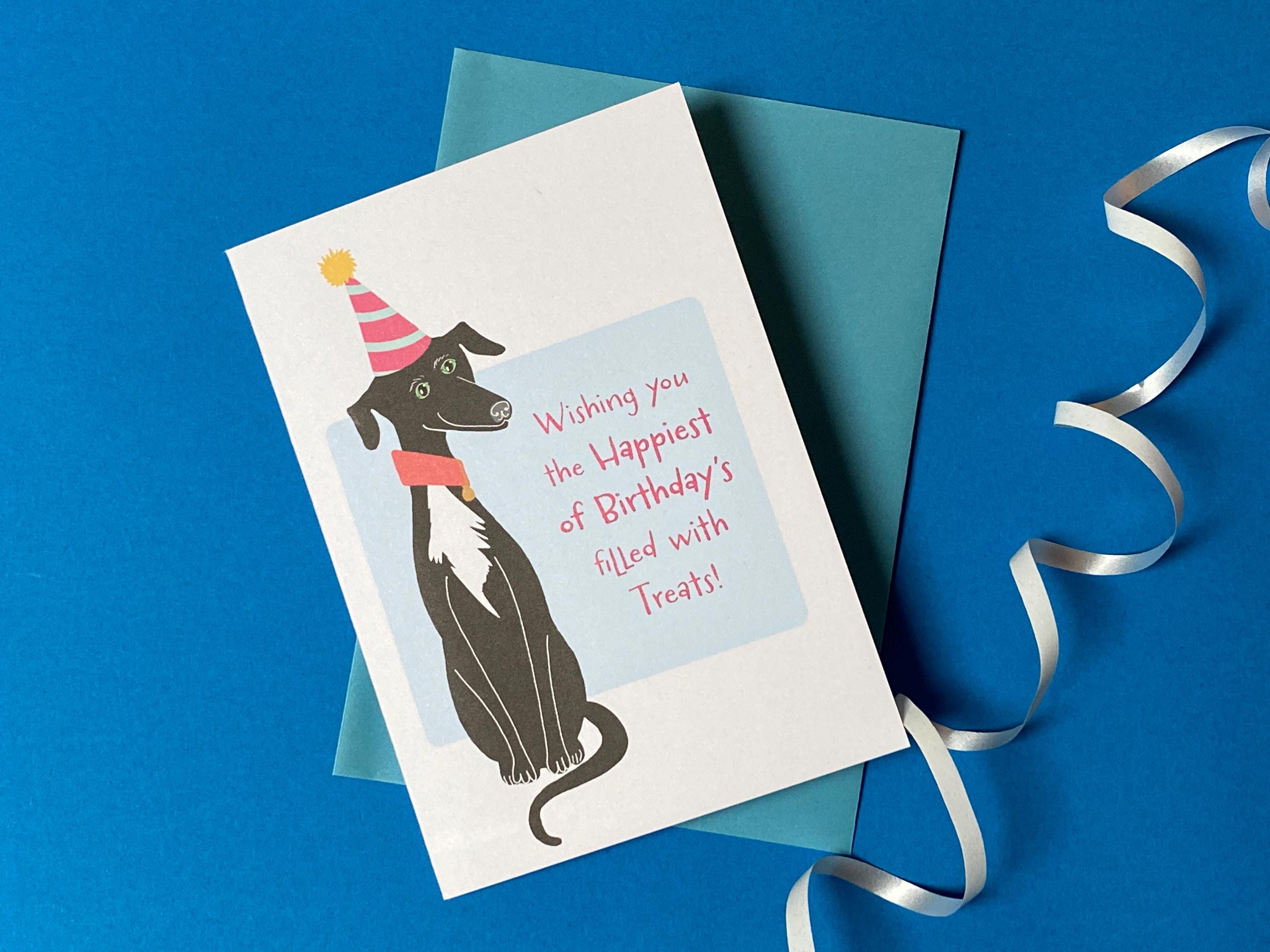 A birthday card sits on a pale blue envelope on a rich blue background with a curly ribbon. The birthday card has a illustration of a seated black greyhound with a white chest and stripy cone shaped birthday hat. The message on the front reads - Wishing you the happiest of Birthdays filled with treats. The text is in pink on a very light blue background.