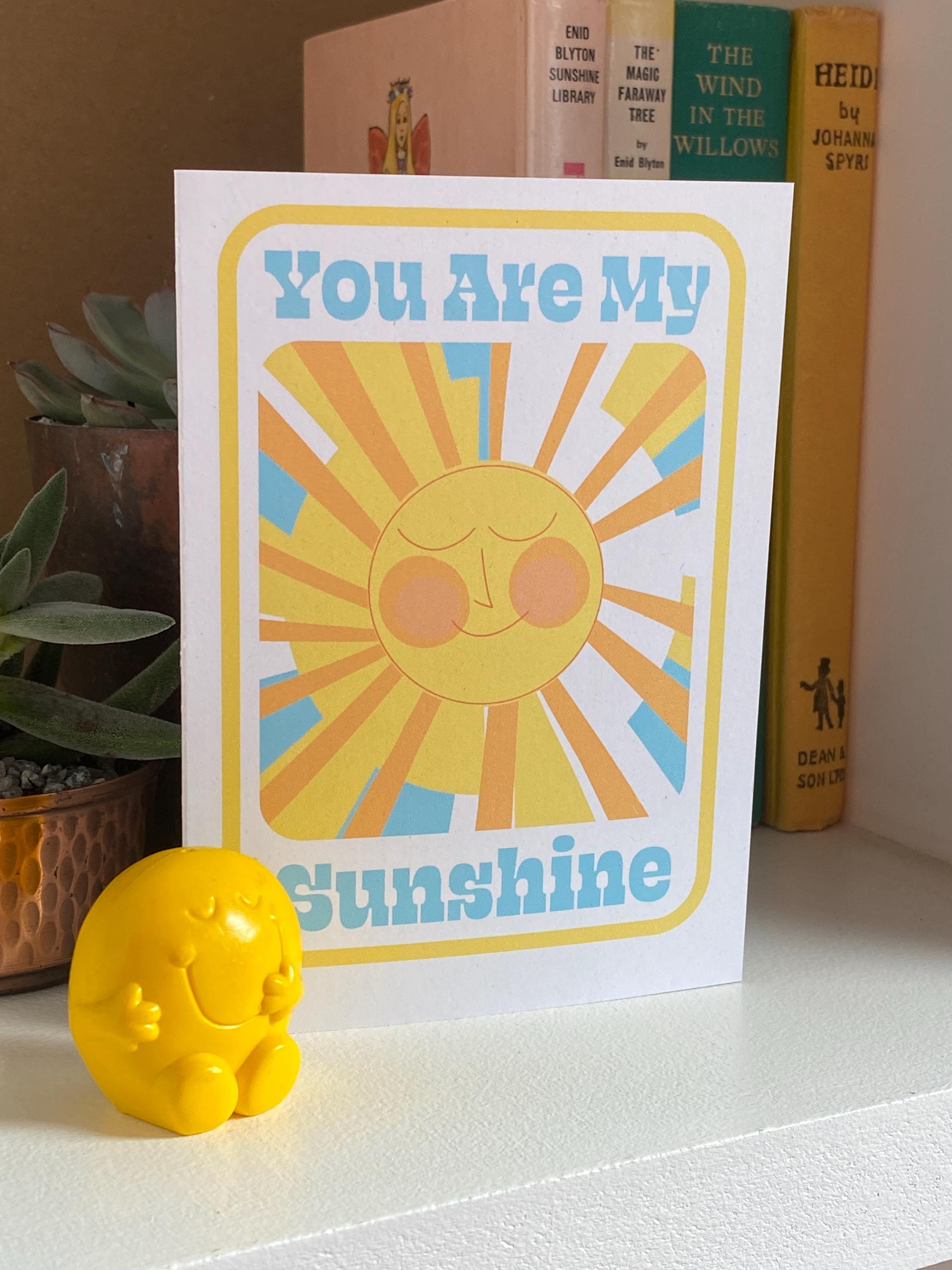 A side view of a birthday card sitting on on a shelf with three books and a plant in the background. The Birthday card is white with smiley illustrated sun in yellows and oranges with rays shining out from it. The text on the card reads You are my sunshine in light blue.