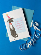 A birthday card sits on a pale blue envelope on a rich blue background with a curly ribbon. The birthday card has a illustration of a seated black greyhound with a white chest and stripy cone shaped birthday hat. The message on the front reads - Wishing you the happiest of Birthdays filled with treats. The text is in pink on a very light blue background.