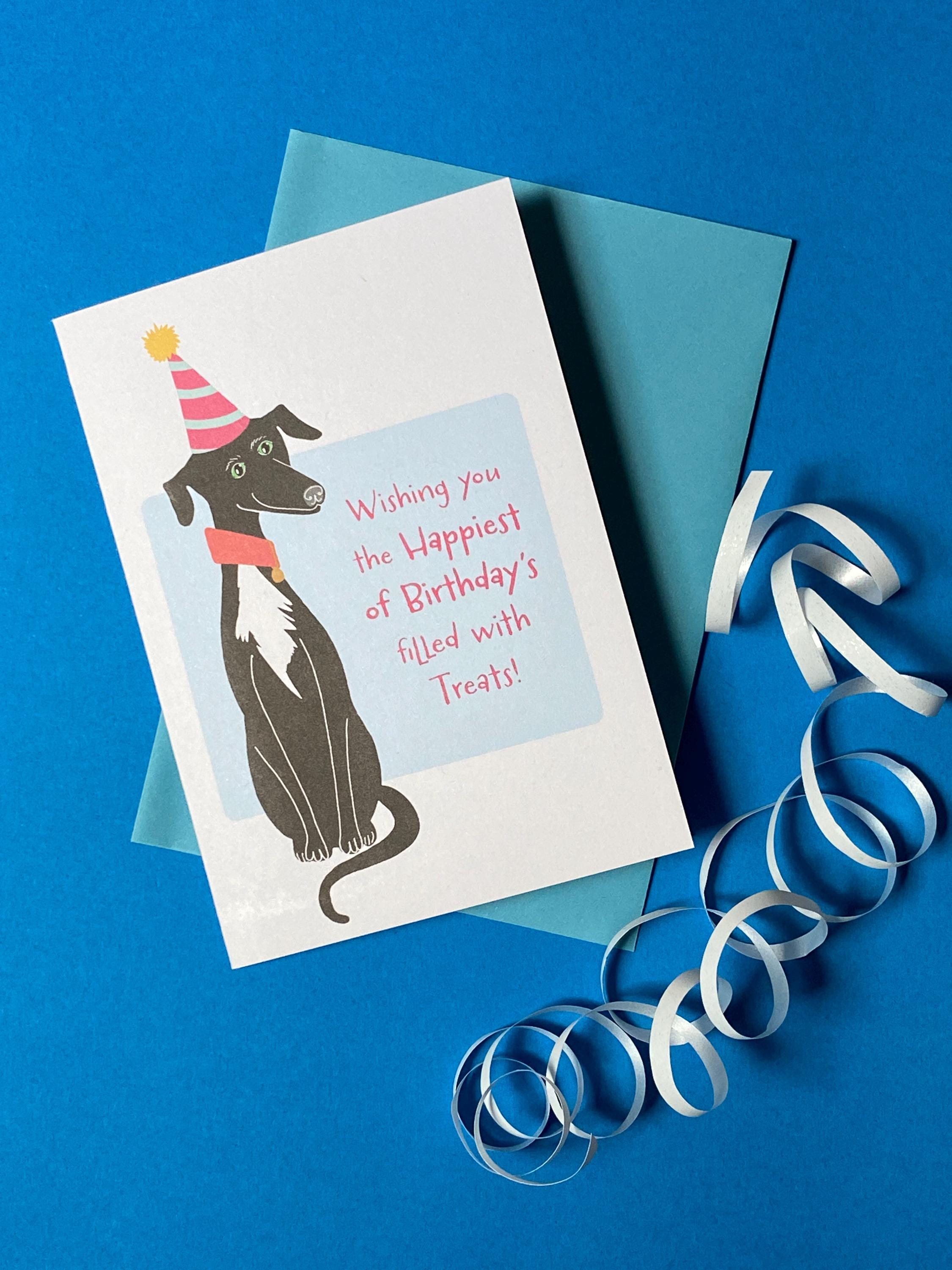 A birthday card sits on a pale blue envelope on a rich blue background with a curly ribbon. The birthday card has a illustration of a seated black greyhound with a white chest and stripy cone shaped birthday hat. The message on the front reads - Wishing you the happiest of Birthdays filled with treats. The text is in pink on a very light blue background.