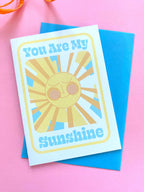 A birthday card sits on a blue envelope on a pink background with an orange ribbon at the side. The Birthday card is white with smiley illustrated sun in yellows and oranges with rays shining out from it. The text on the card reads You are my sunshine in light blue.