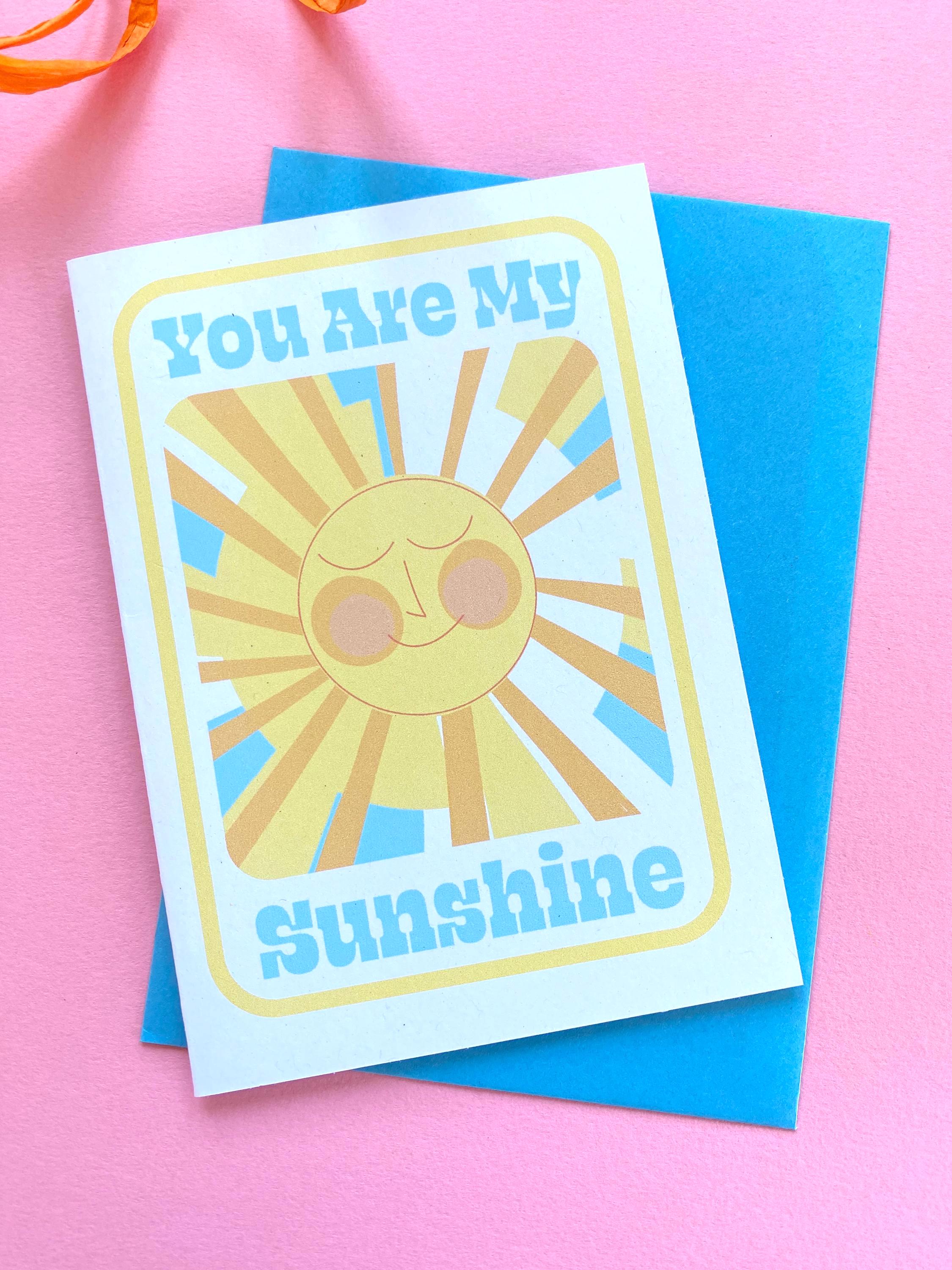 A birthday card sits on a blue envelope on a pink background with an orange ribbon at the side. The Birthday card is white with smiley illustrated sun in yellows and oranges with rays shining out from it. The text on the card reads You are my sunshine in light blue.