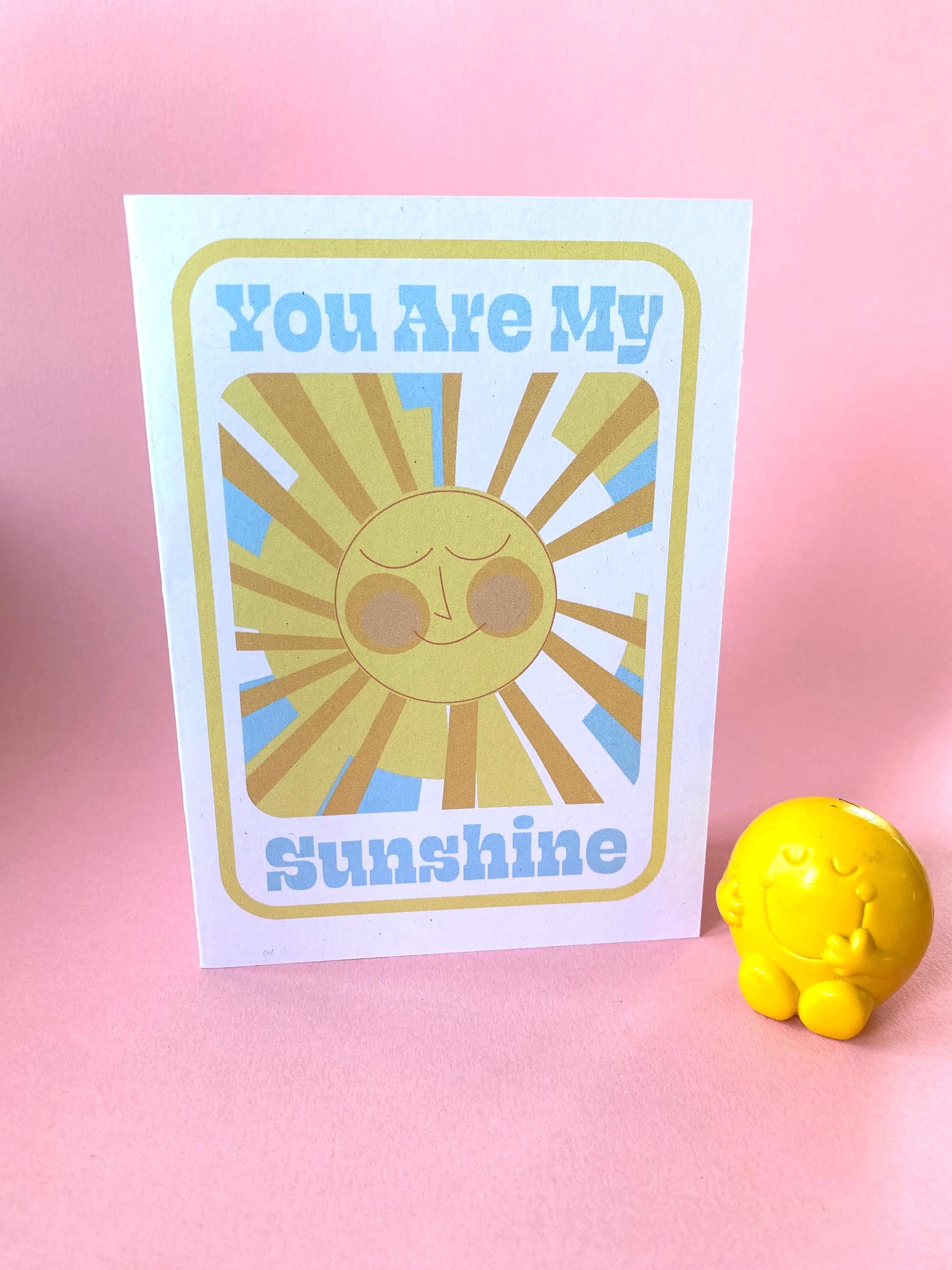 A birthday card stands on a pink background with yellow character at the side. The Birthday card is white with smiley illustrated sun in yellows and oranges with rays shining out from it. The text on the card reads You are my sunshine in light blue.