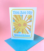 A birthday card stands on a blue envelope on a pink background. The Birthday card is white with smiley illustrated sun in yellows and oranges with rays shining out from it. The text on the card reads You are my sunshine in light blue.