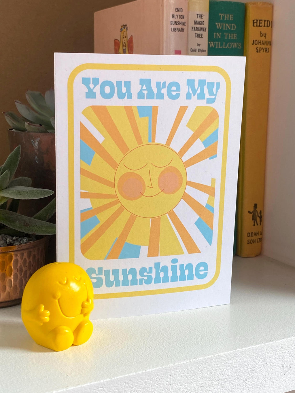 A birthday card sits on on a shelf with three books and a plant in the background. The Birthday card is white with smiley illustrated sun in yellows and oranges with rays shining out from it. The text on the card reads You are my sunshine in light blue.