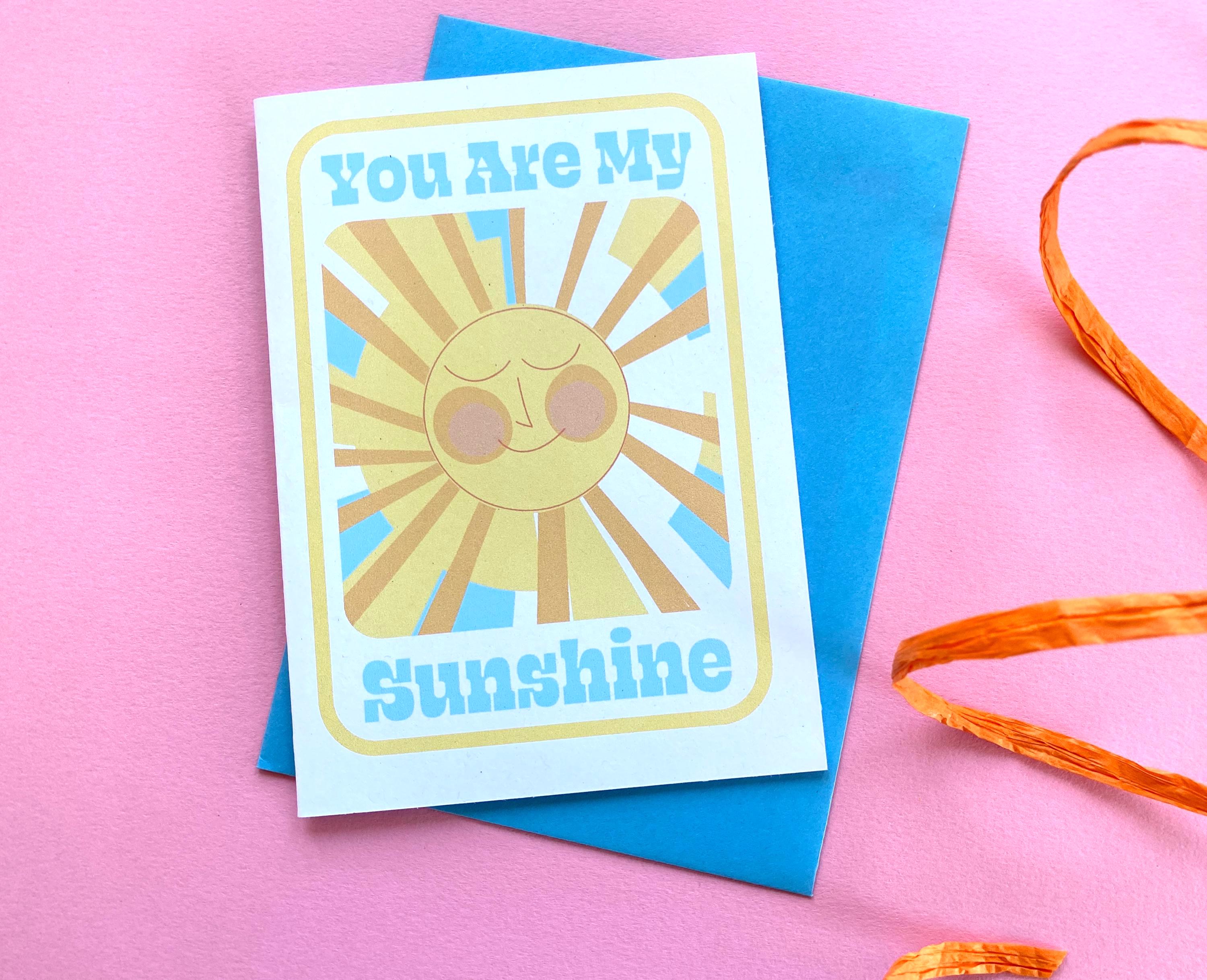 A birthday card sits on a blue envelope on a pink background with an orange ribbon at the side. The Birthday card is white with smiley illustrated sun in yellows and oranges with rays shining out from it. The text on the card reads You are my sunshine in light blue.