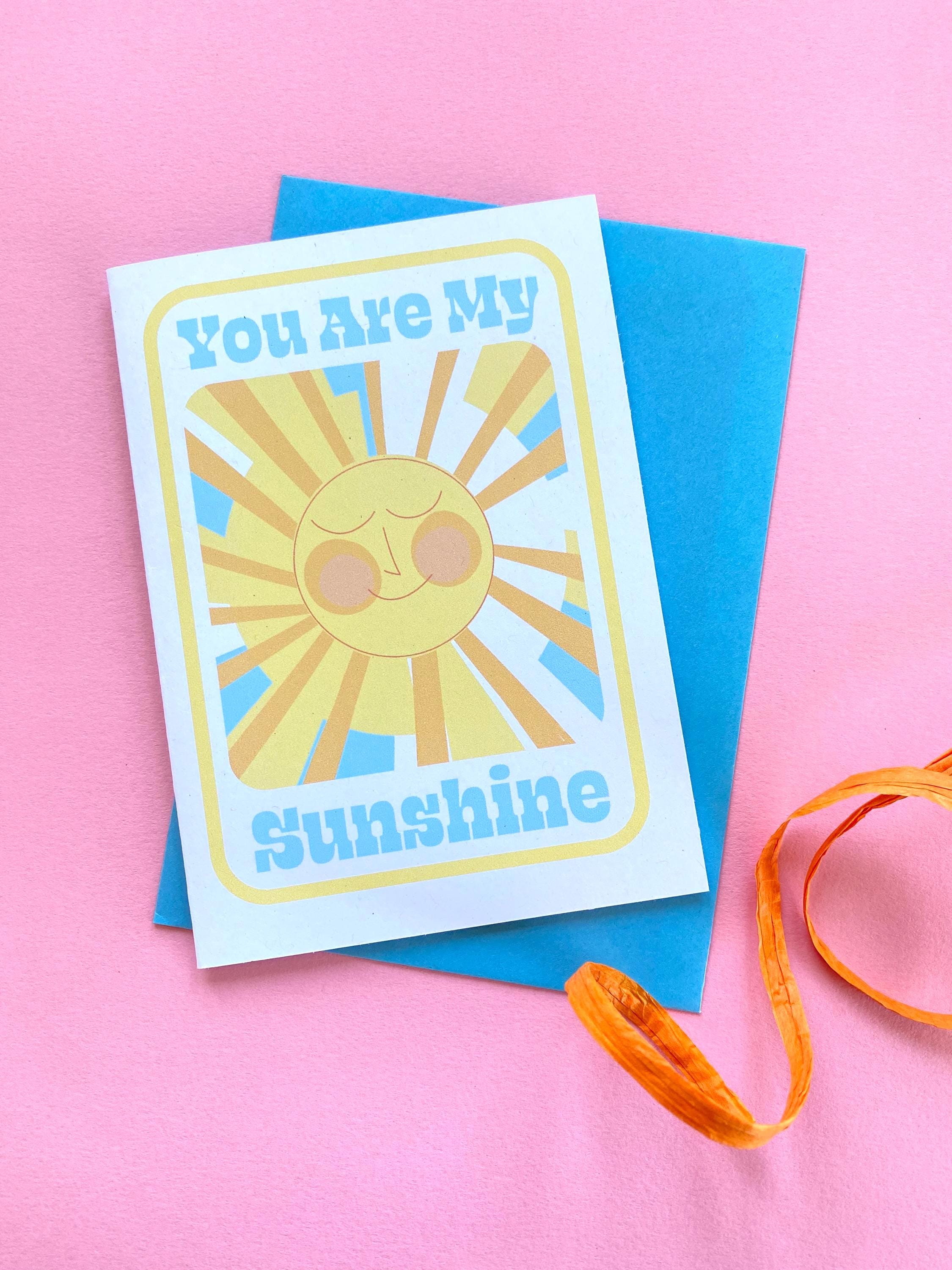 A birthday card sits on a blue envelope on a pink background with an orange ribbon at the side. The Birthday card is white with smiley illustrated sun in yellows and oranges with rays shining out from it. The text on the card reads You are my sunshine in light blue.