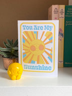 A birthday card sits on on a shelf with three books and a plant in the background. The Birthday card is white with smiley illustrated sun in yellows and oranges with rays shining out from it. The text on the card reads You are my sunshine in light blue.